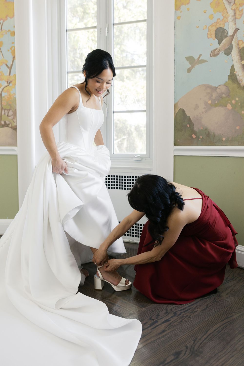 A helper in burgundy assists with the bride's shoes during wedding preparations in a decorative room.
