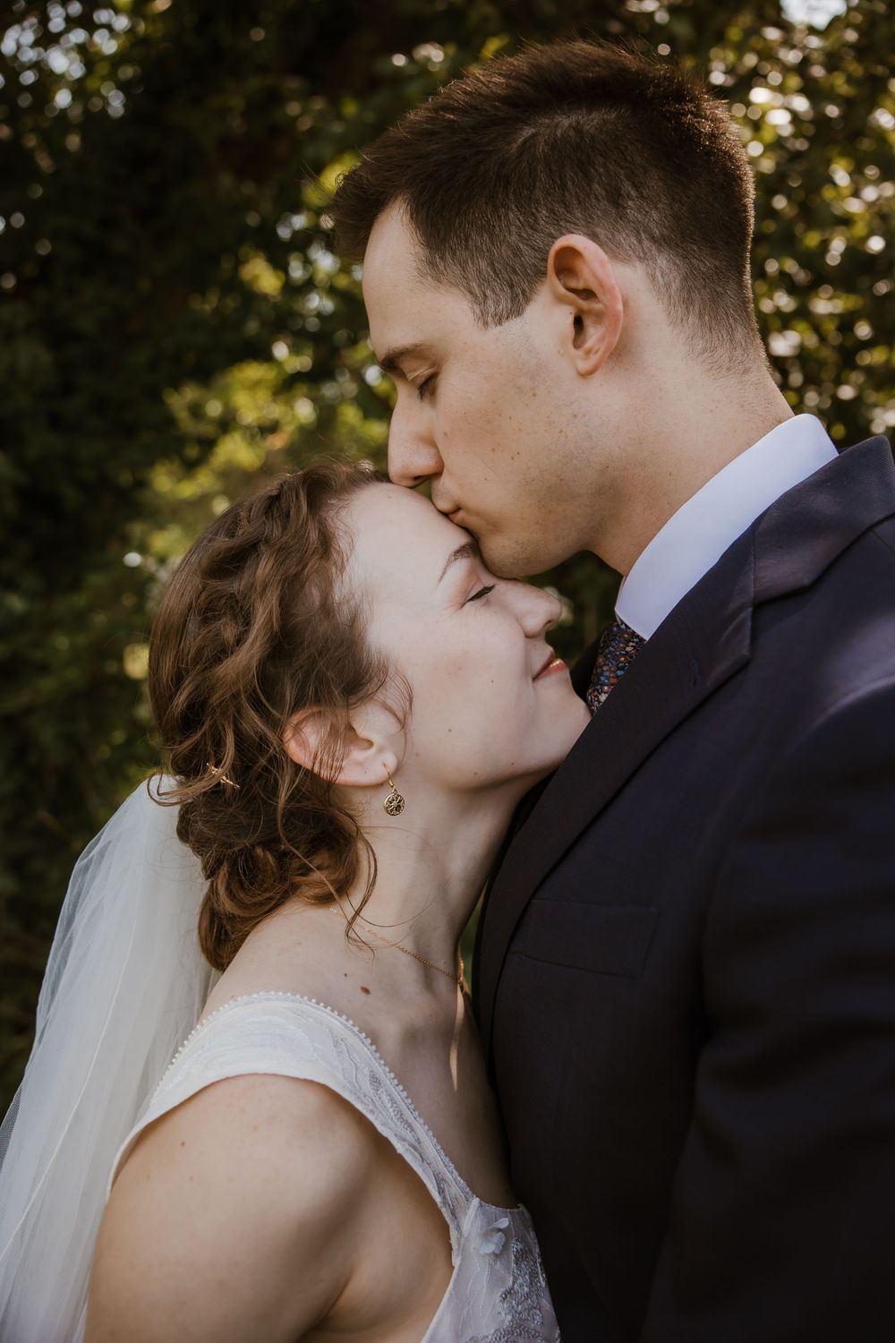 A romantic wedding moment captured outdoors with the groom kissing the bride's forehead in natural lighting.