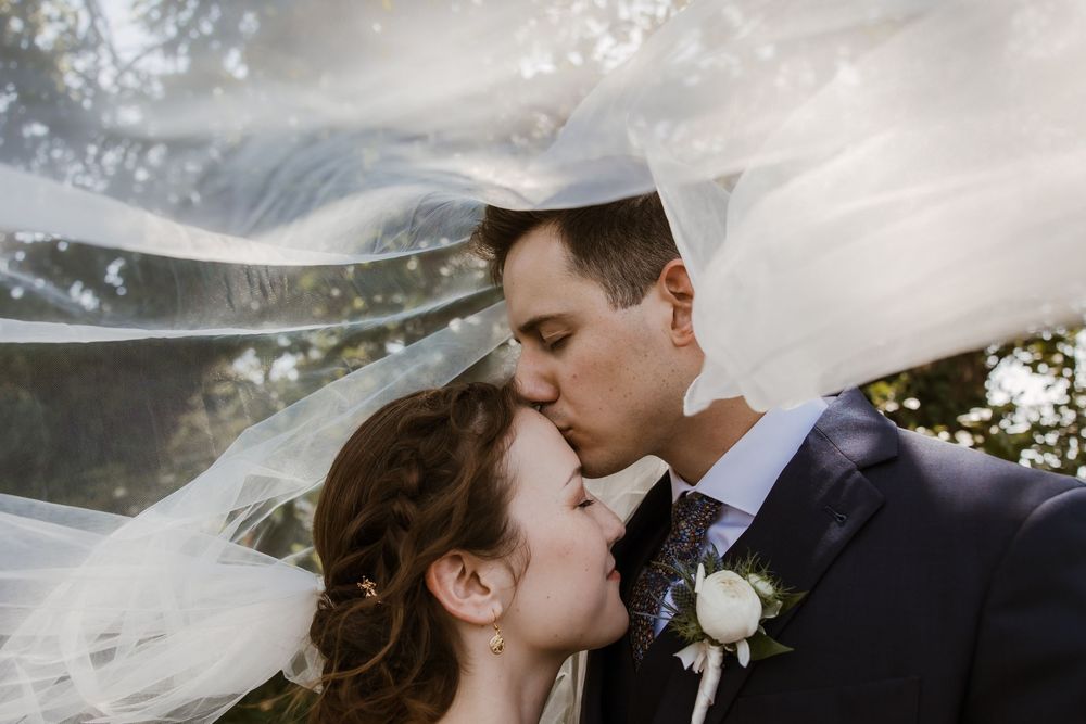 A romantic wedding moment captured under a flowing veil as a couple shares a tender kiss outdoors.