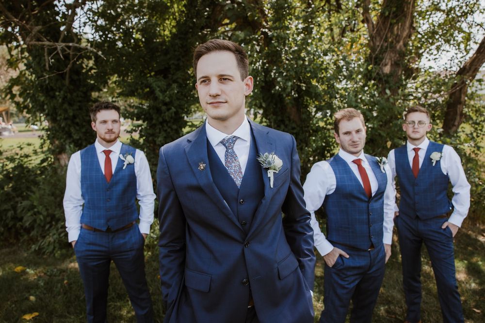 Groomsmen in navy blue suits and red vests pose outdoors in a natural setting for a wedding party photo. Kansas City Wedding Photography