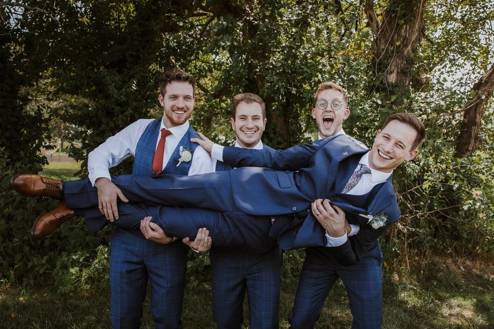 Groomsmen in navy blue suits laughing together while carrying groom horizontally and bouncing him. Meadowbrook Park, Kansas City wedding day