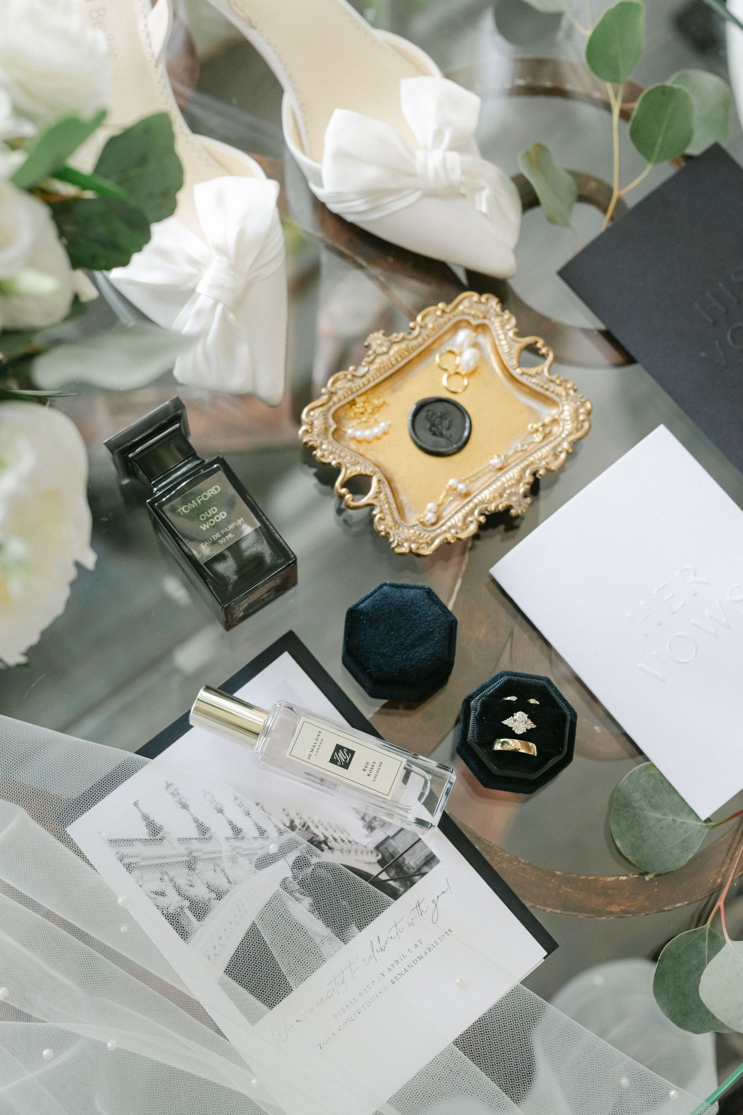 White roses and eucalyptus in mason jar with wedding accessories and vow books displayed on glass table.