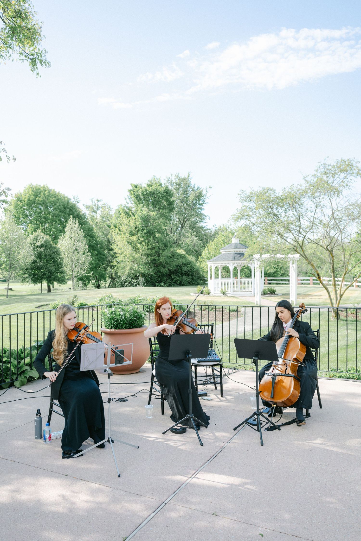 String quartet musicians perform classical music outdoors on a patio with a white gazebo and green landscape in background.