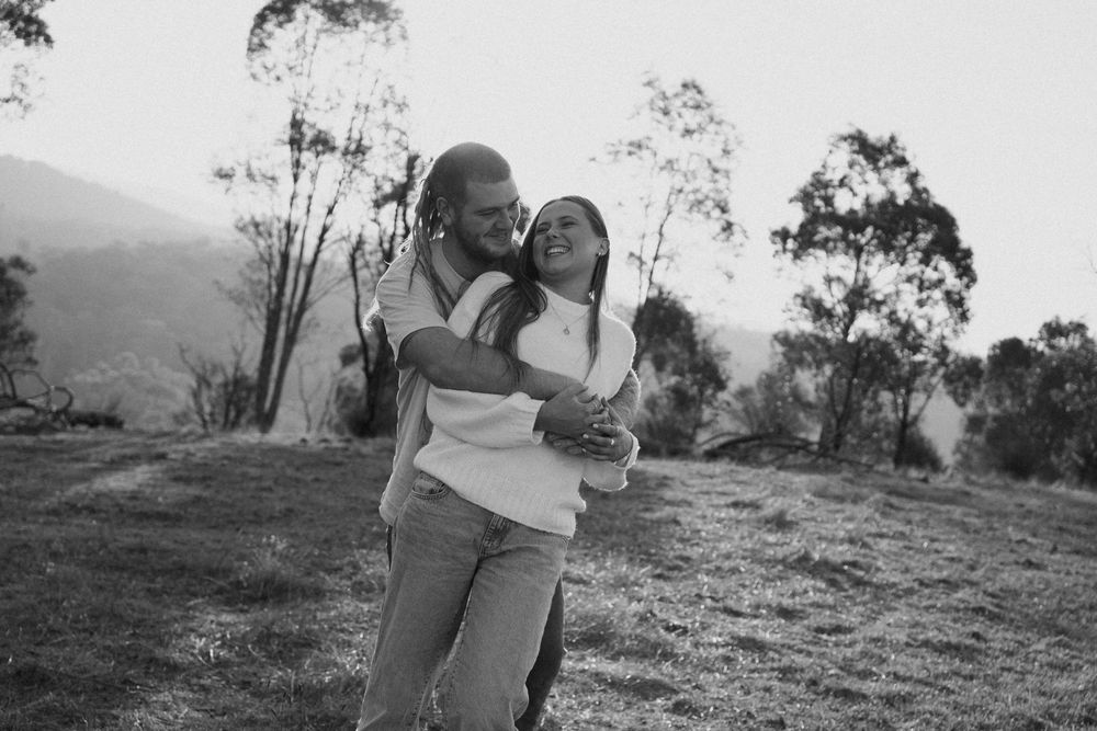A joyful black and white outdoor portrait shows two people embracing while laughing together on a hillside with trees.