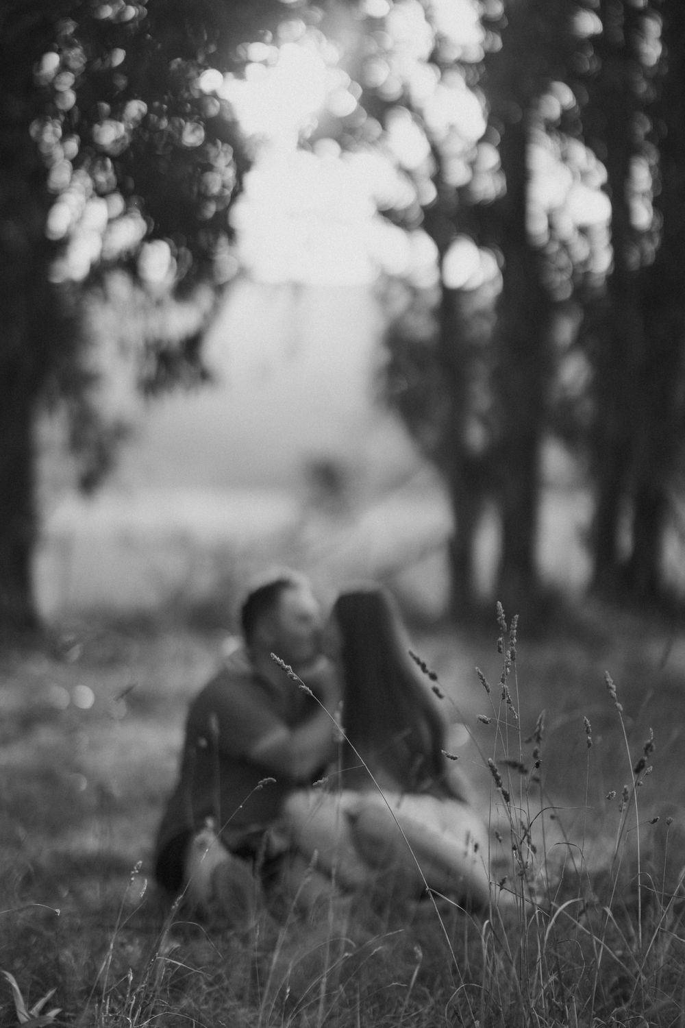 A dreamy black and white photo showing two people sitting together in a meadow with soft bokeh trees in the background.