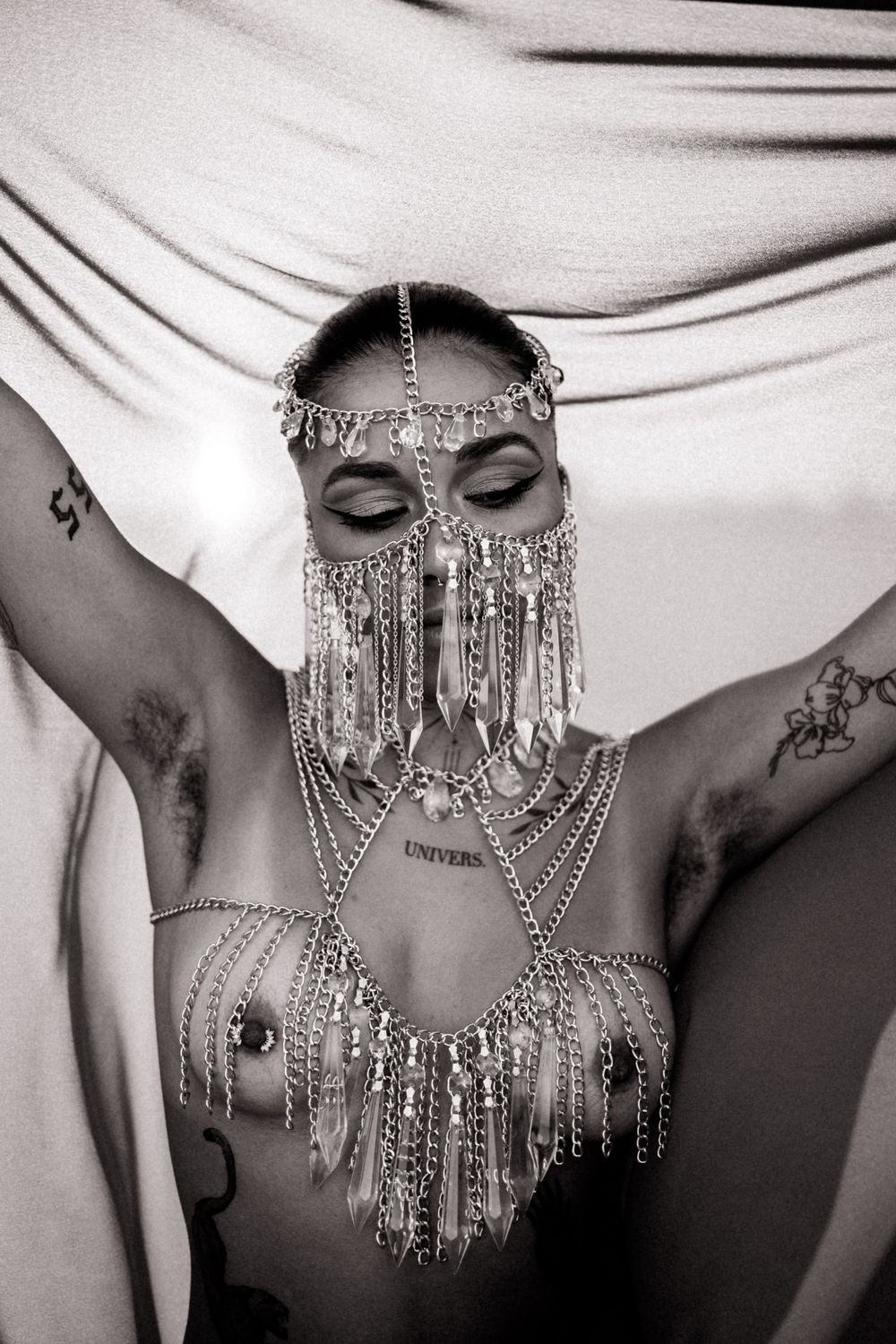 Black and white artistic photo of person wearing intricate beaded face jewelry and body chain with draped fabric backdrop.