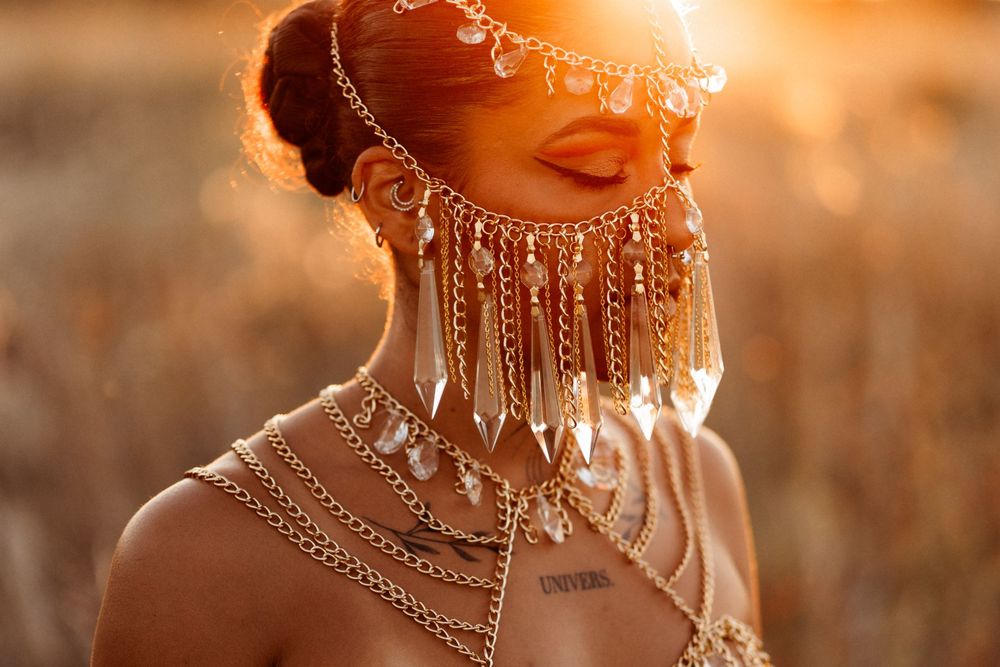 Ornate silver jewelry and chains glow in golden sunlight against golden grass in a dreamlike portrait.