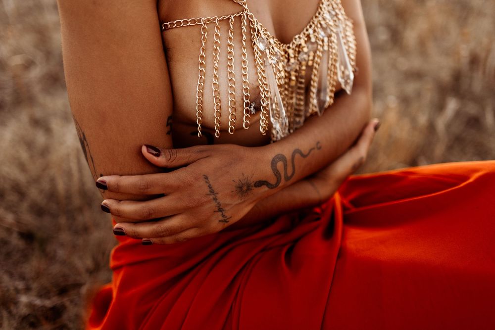 Close up of hands crossed over torso wearing beaded jewelry and red flowing garment against dried grass background.