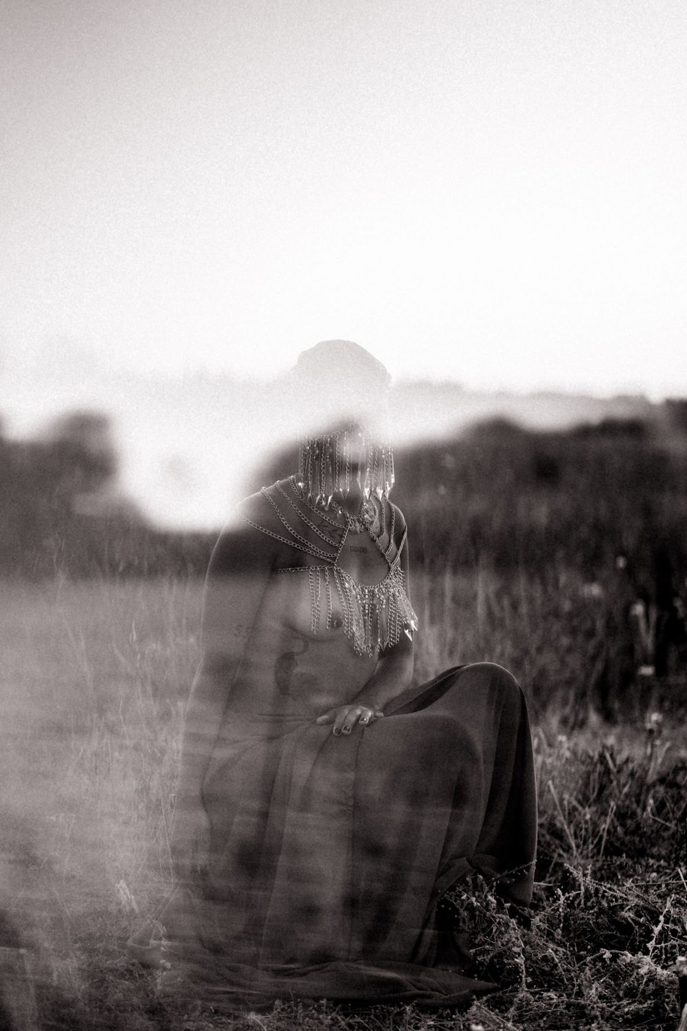 A ghostly double-exposed black and white photograph of a seated figure in a field at sunset.