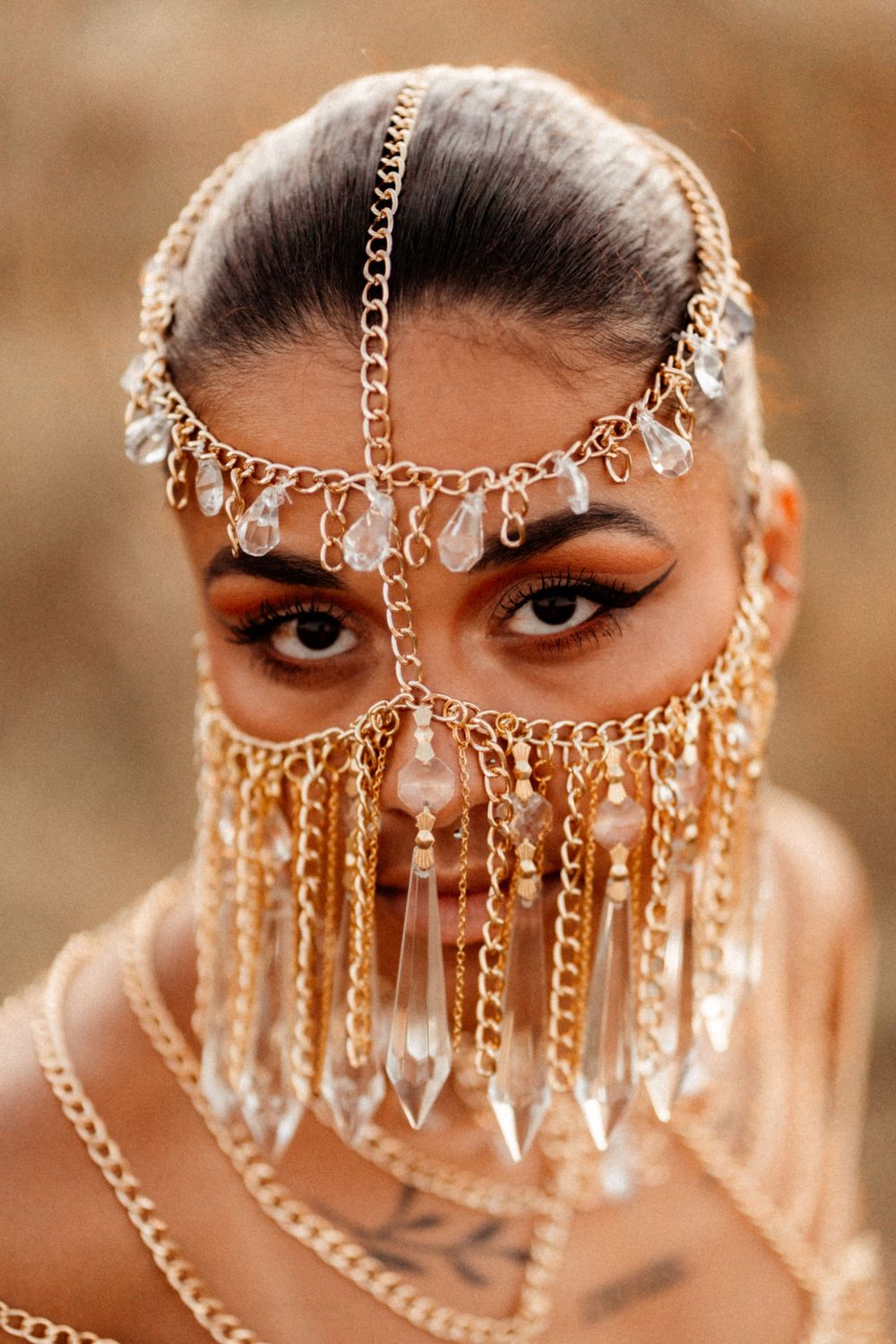 Ornate gold chain headpiece with crystal drops and chains drapes across face with a veil-like effect against warm brown tones.
