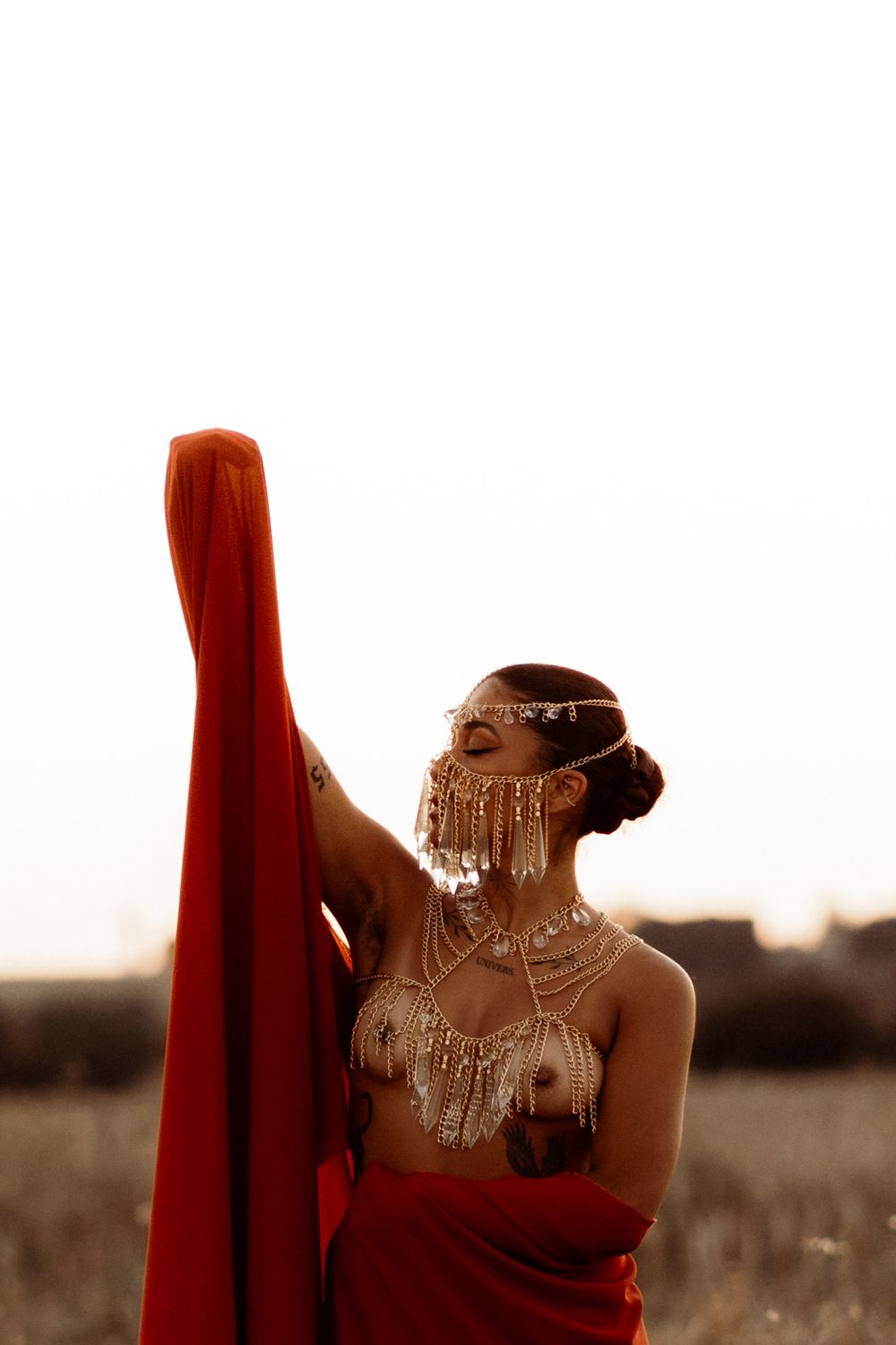 Artistic silhouette of a person in red fabric adorned with tribal jewelry against a sepia-toned landscape at sunset.
