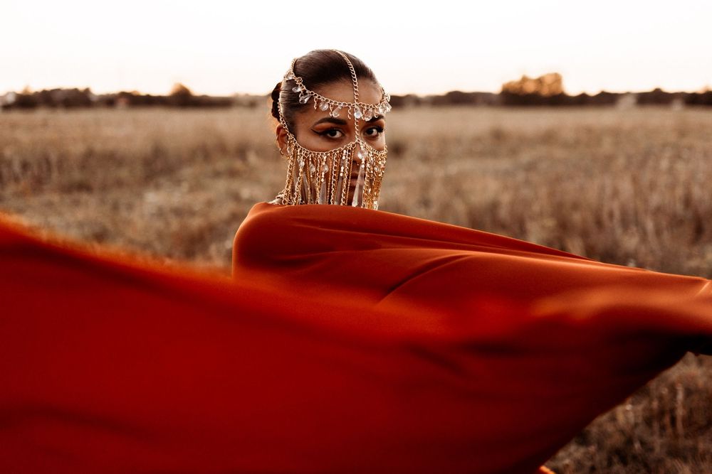 A figure wrapped in flowing red fabric stands in a golden field at sunset with sunglasses on.