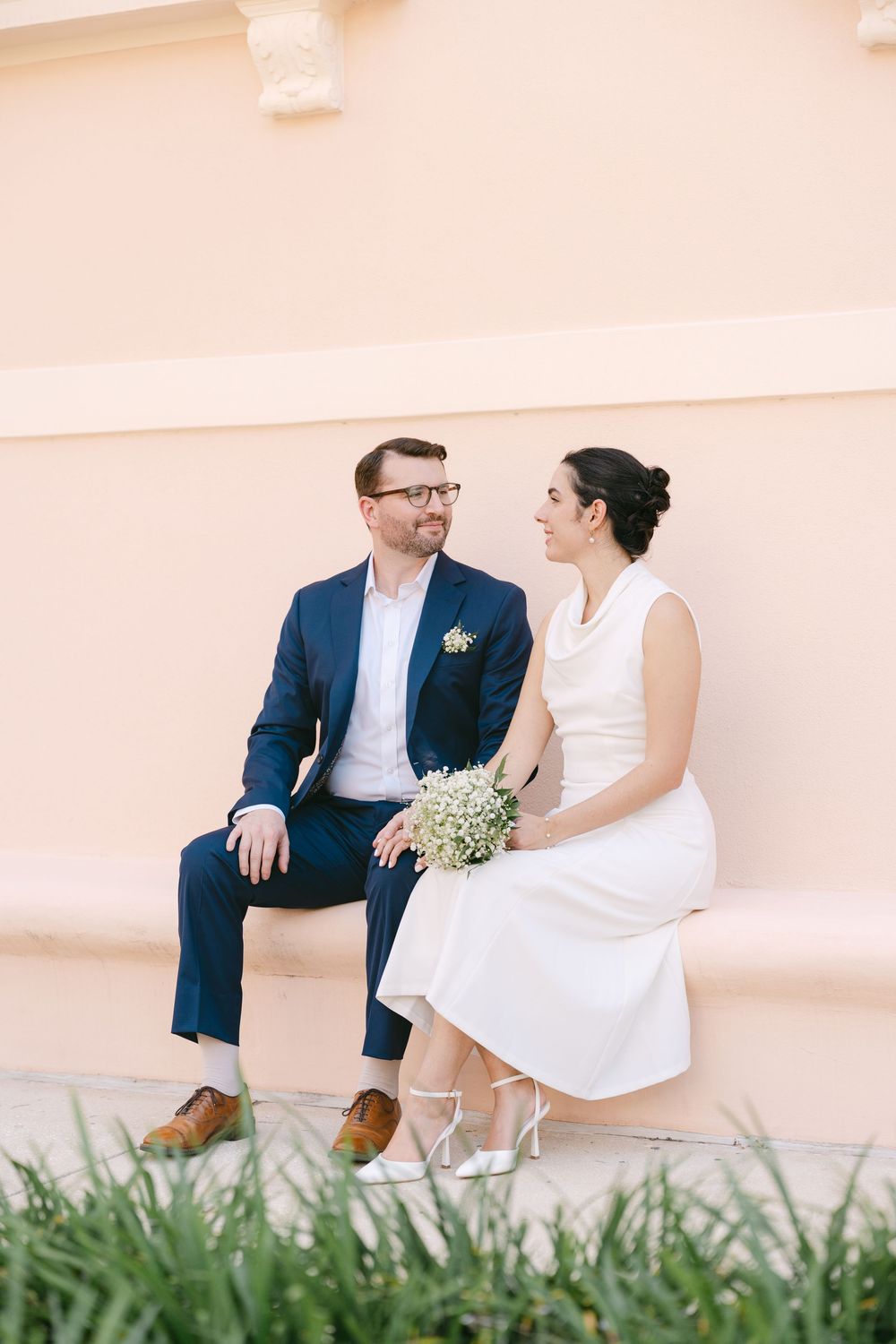 A couple in wedding attire sits together on a pink wall ledge with tropical foliage in the foreground.