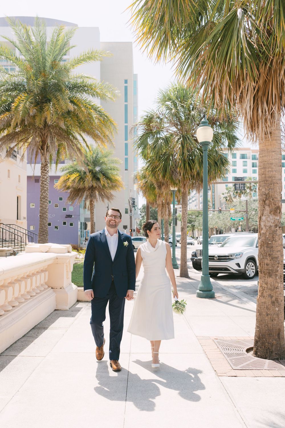A couple walks down a palm tree-lined street in wedding attire on a sunny day.