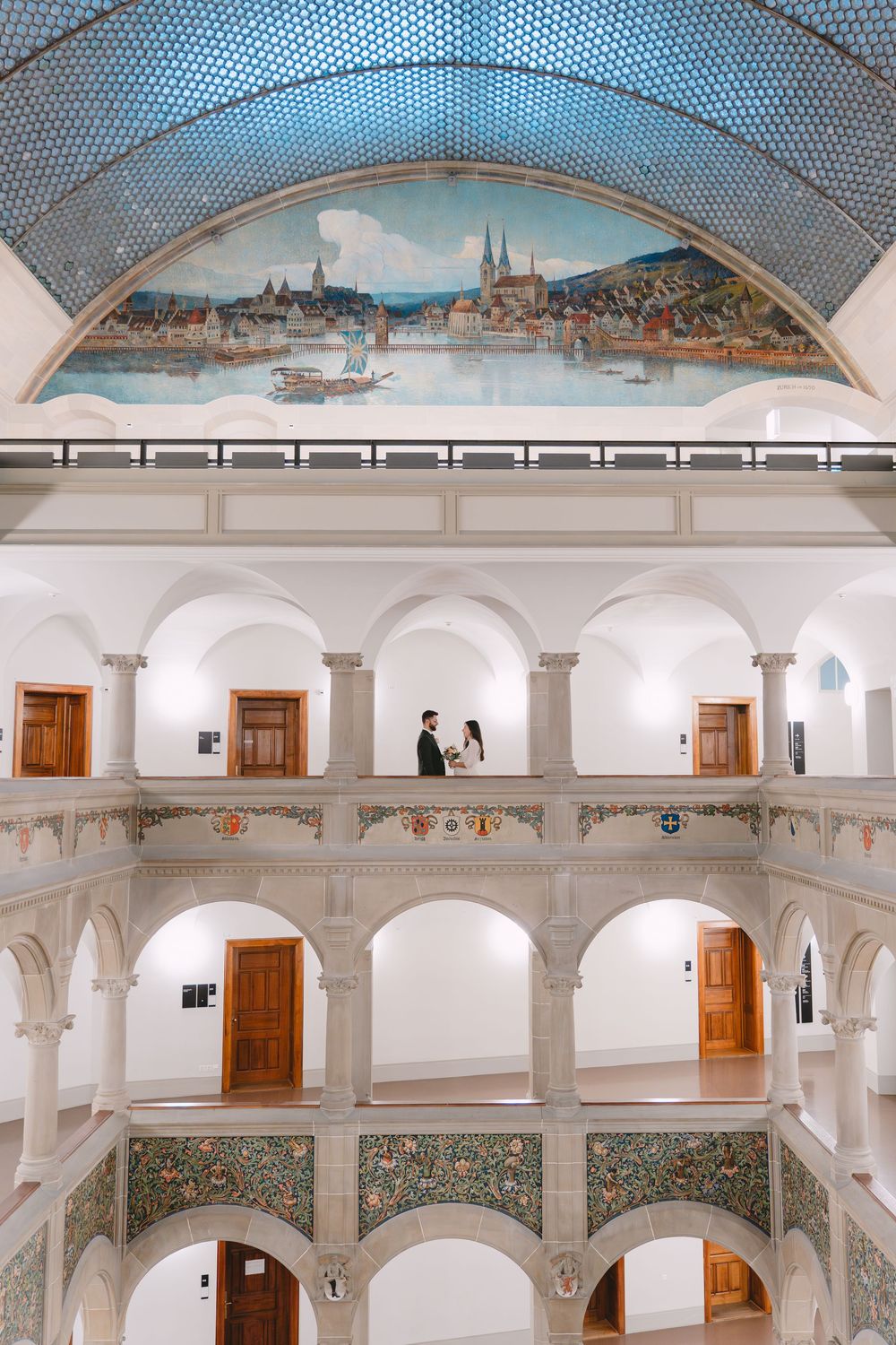 A multi-level interior atrium with white arches and wooden doors features a scenic painted mural on the vaulted ceiling.