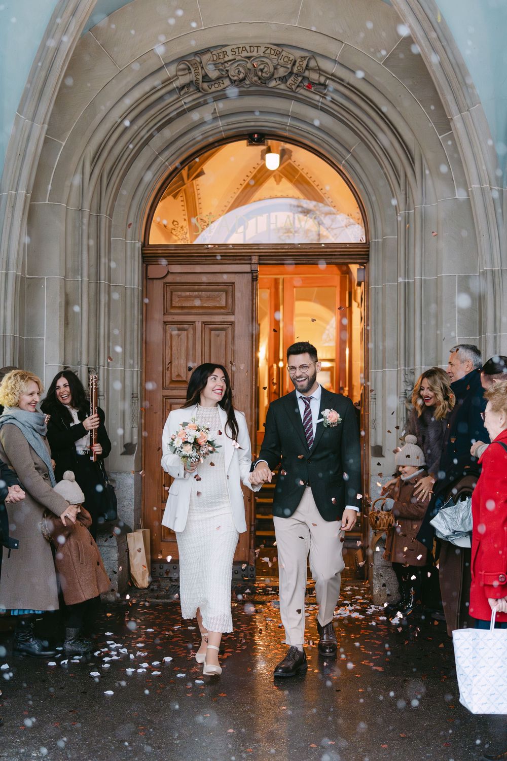 Couple exits church through stone archway while guests toss confetti during winter wedding celebration.