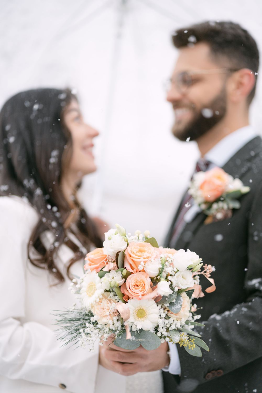 Romantic winter wedding couple gazes at each other while holding a blush pink and white floral bouquet in the falling snow.