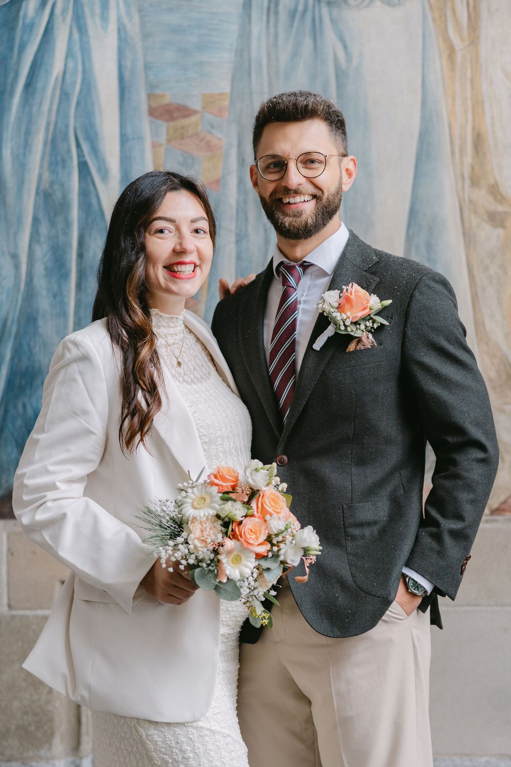 A couple poses at their wedding with a beautiful peach and coral floral bouquet against a light blue backdrop.