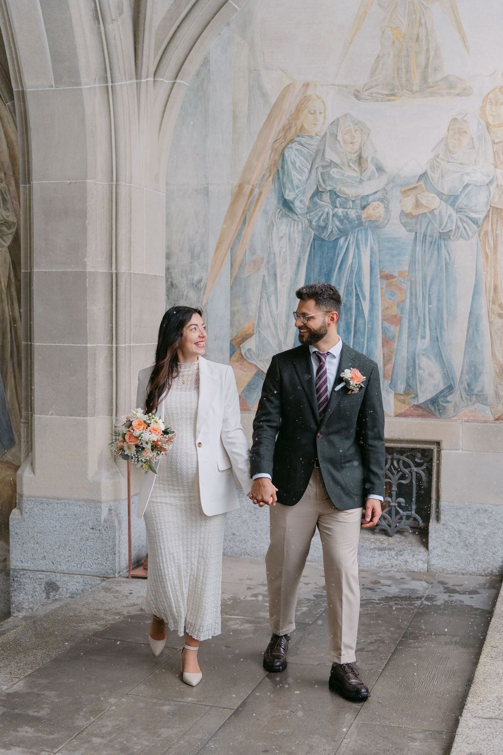 A couple walking together in formal attire along an ornate church corridor with religious murals on the wall.