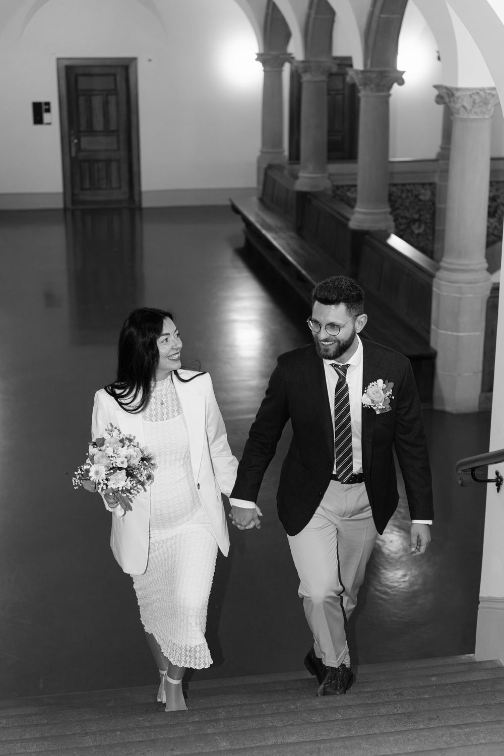 A couple walks hand in hand through a corridor with arched columns, the bride wearing a modern white pantsuit.