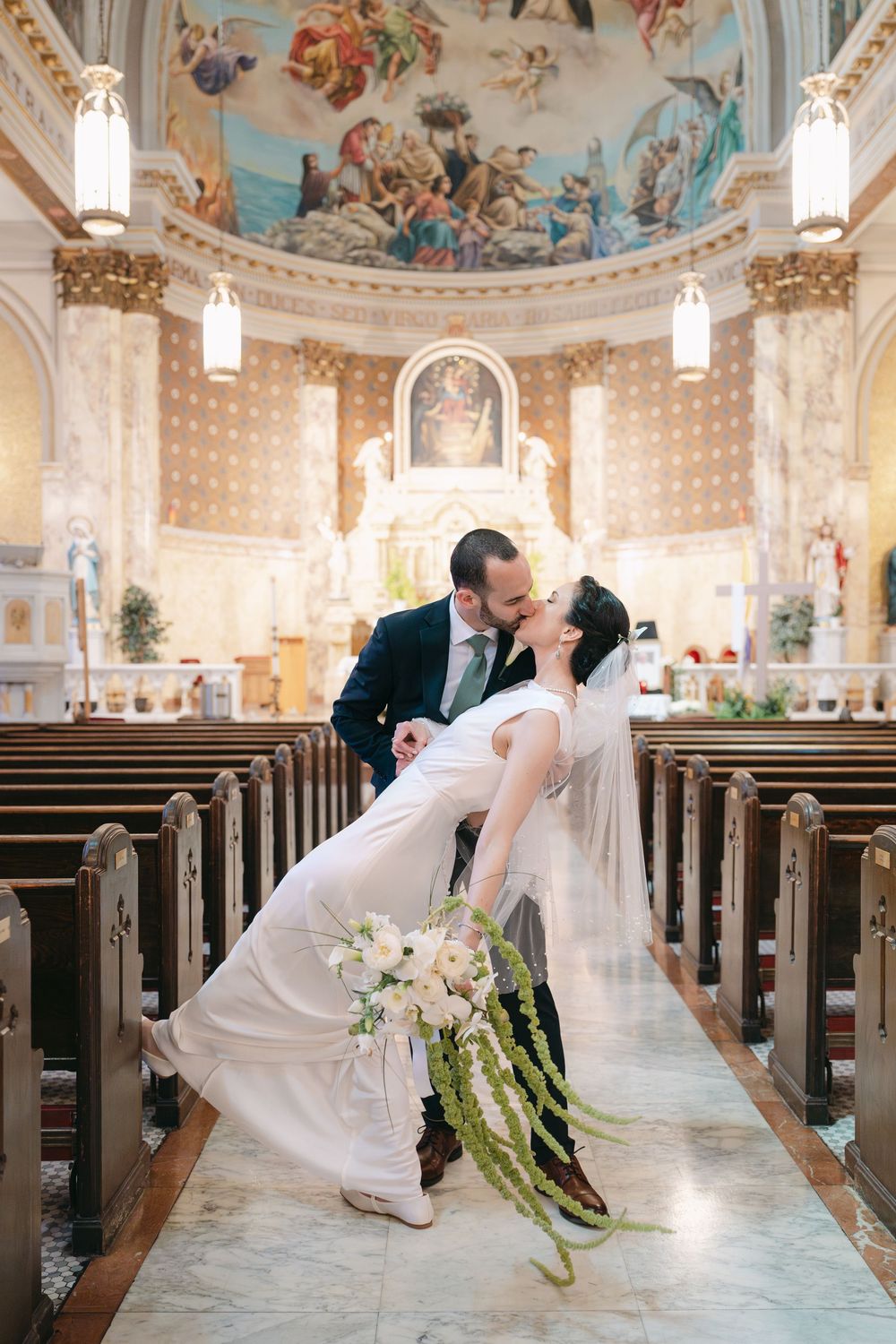 A couple shares a romantic dip kiss in an ornate church aisle with a beautiful painted dome ceiling and wooden pews.
