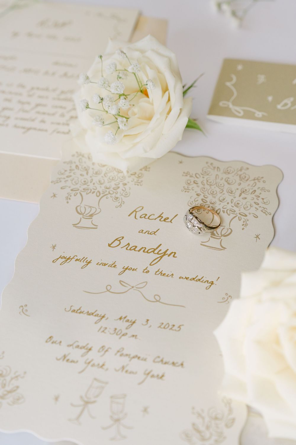 Cream colored wedding invitation with gold calligraphy text and white rose detail against white background.