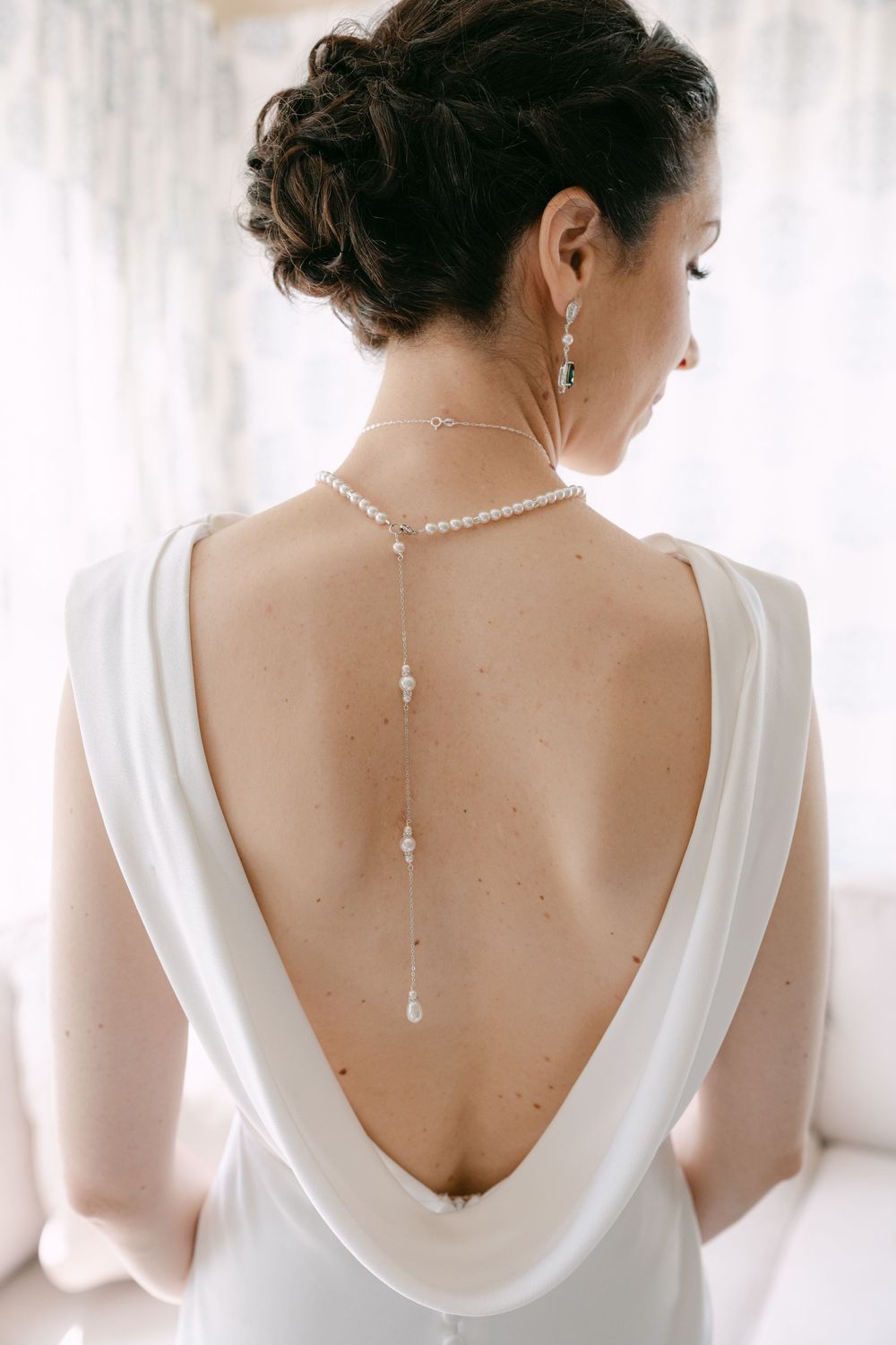 Elegant back necklace adorns a plunging white wedding dress with draped cowl back detail.