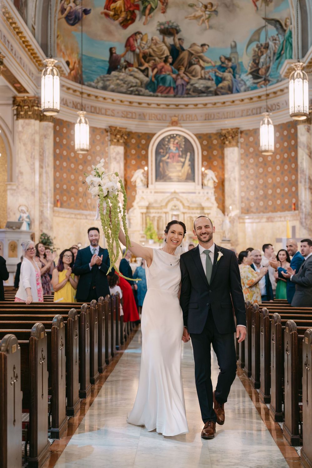 A wedding ceremony exits down the church aisle with ornate murals and hanging lanterns decorating the beautiful sanctuary.