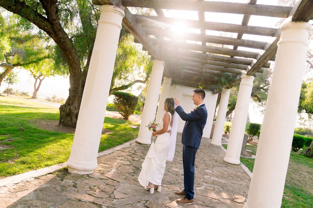 Couple dancing together under a white columned pergola in a sunlit garden setting.