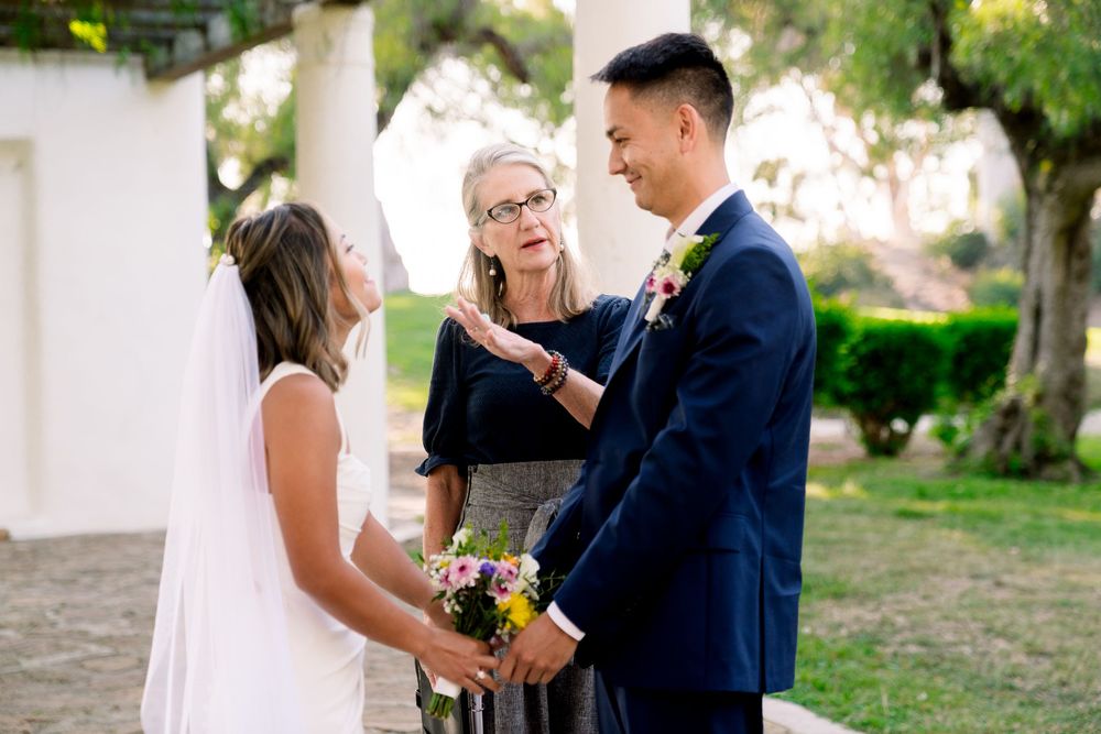 An outdoor wedding ceremony takes place in a garden setting with the officiant presiding over the couple's vows.