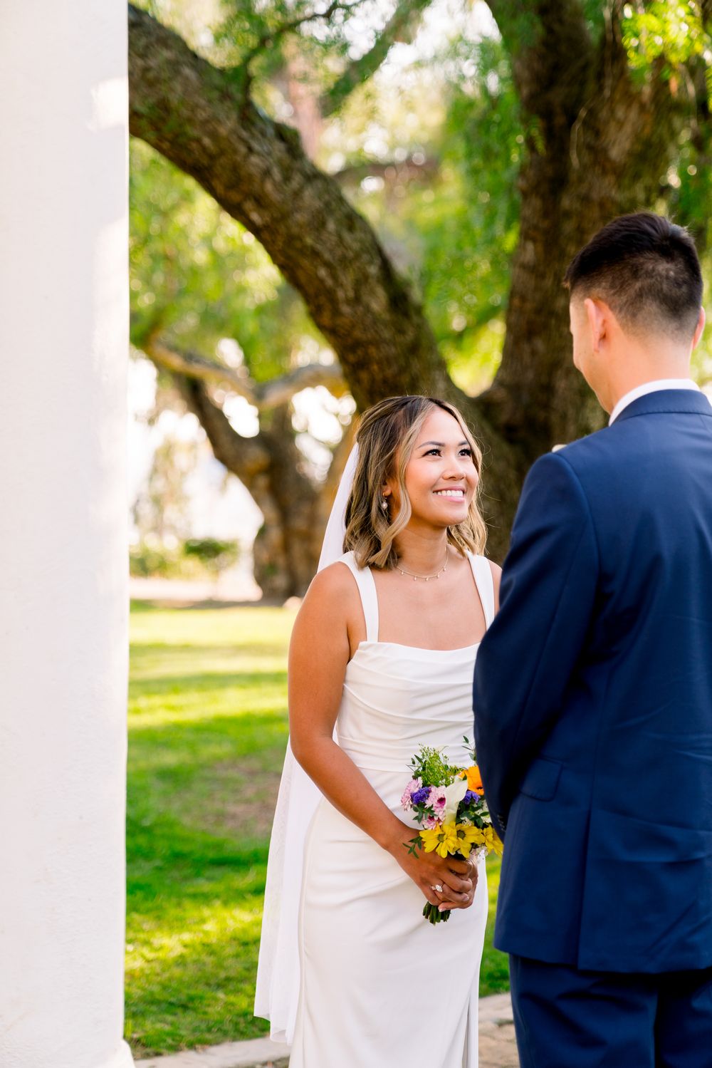 Joyful bride holding a colorful bouquet smiles at her groom during their outdoor wedding ceremony under a large tree.
