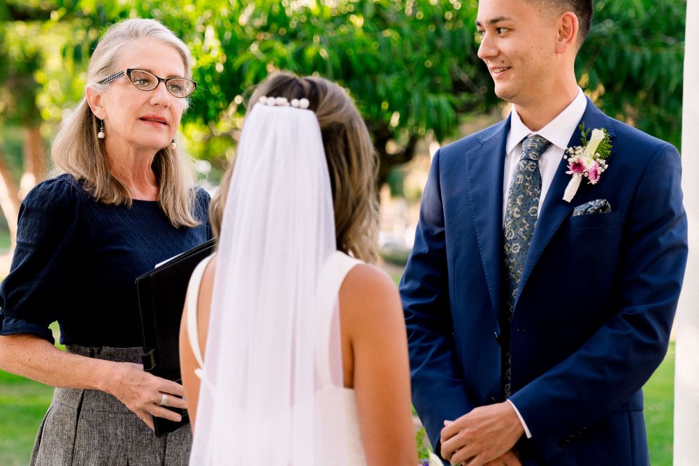 An outdoor wedding ceremony takes place in a garden setting with guests and wedding party members present.