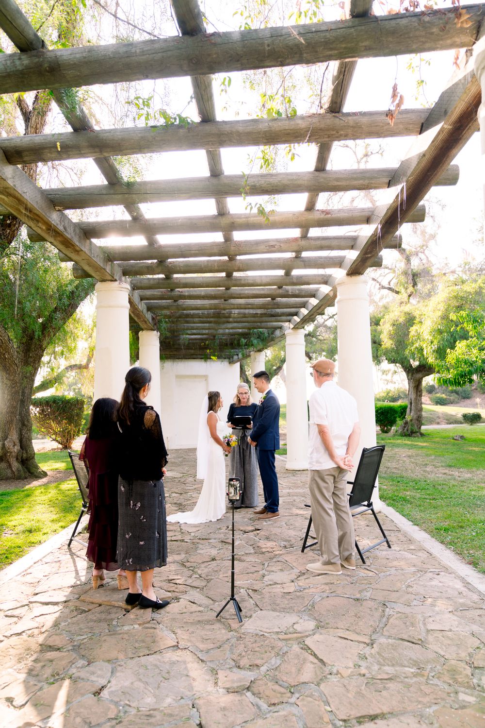 Small outdoor ceremony taking place under rustic wooden pergola on stone patio surrounded by garden scenery.
