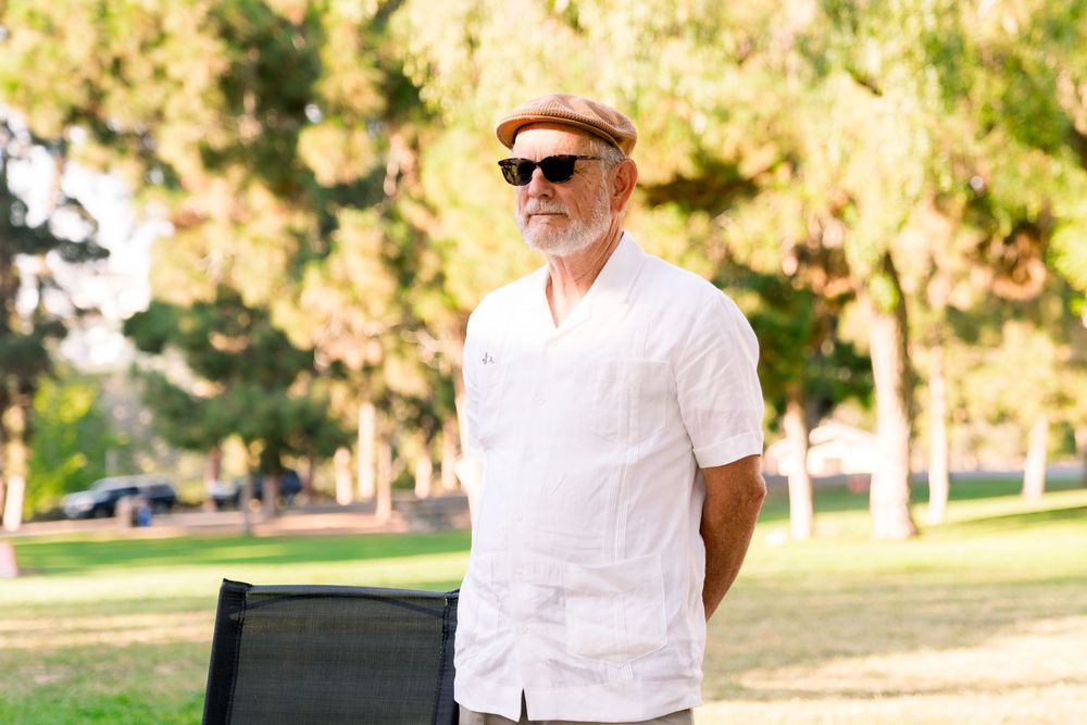 Someone in sunglasses and a white button-down shirt stands confidently in a sunny park setting.