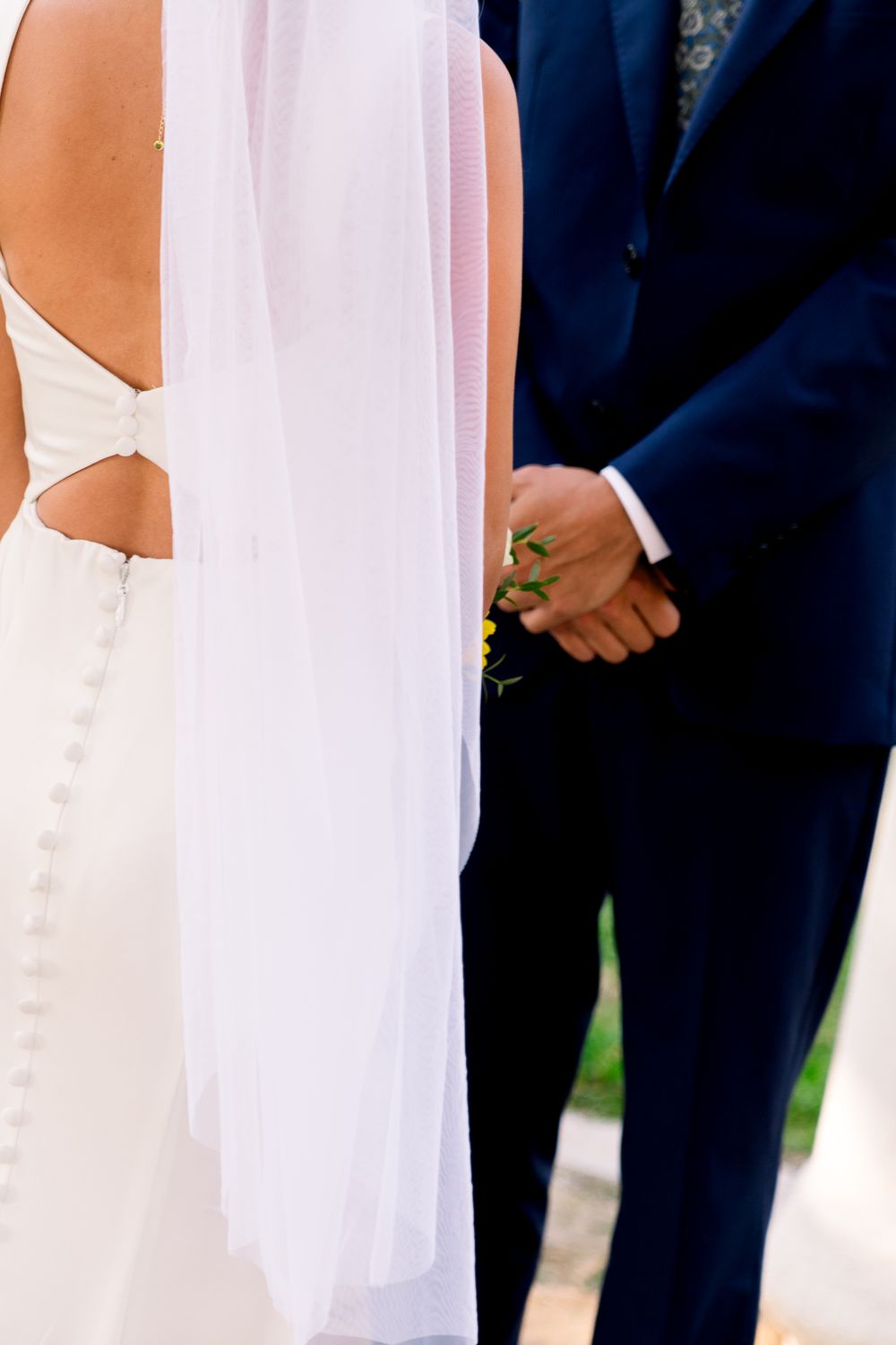 Close-up detail of a bride in white gown with cut-out details next to groom wearing navy suit at wedding ceremony.