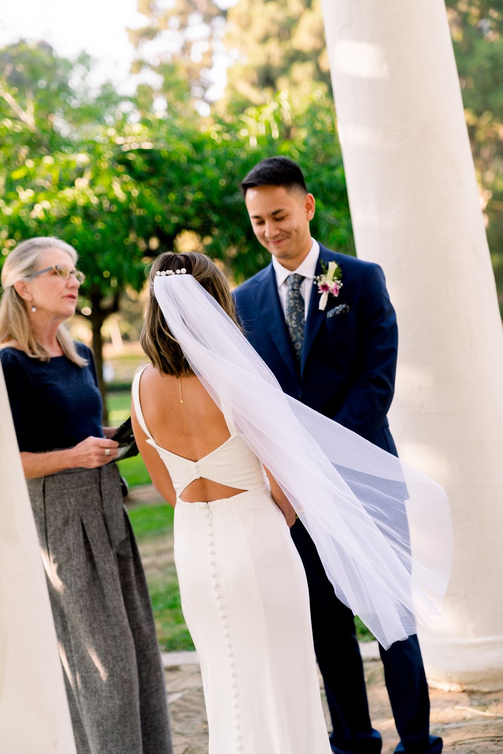 A wedding ceremony takes place outdoors with an elegant white column and lush greenery in the background.