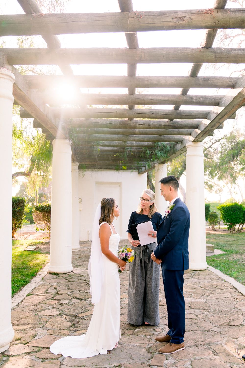 Wedding ceremony takes place under a white columned pergola in a sunlit garden setting.