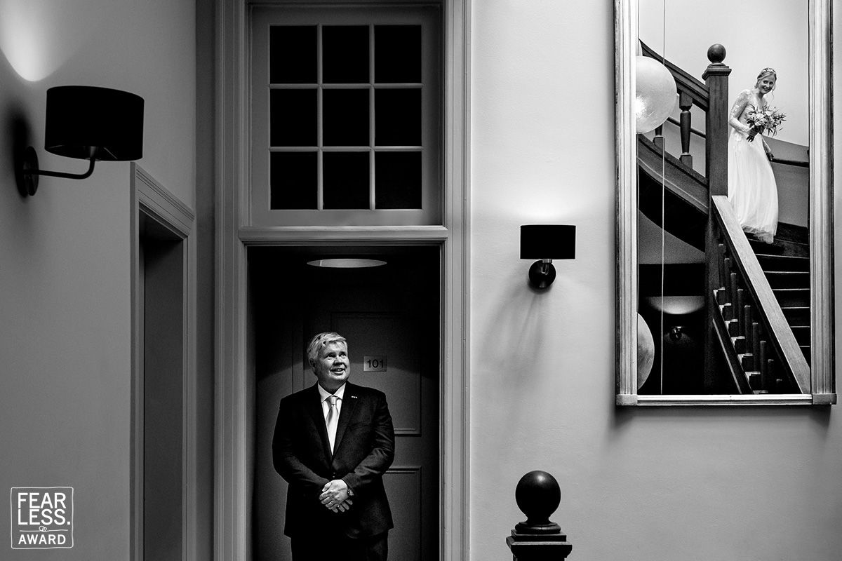 Black and white photo showing someone standing in an elegant doorway under a window with wall sconces and stairs visible.