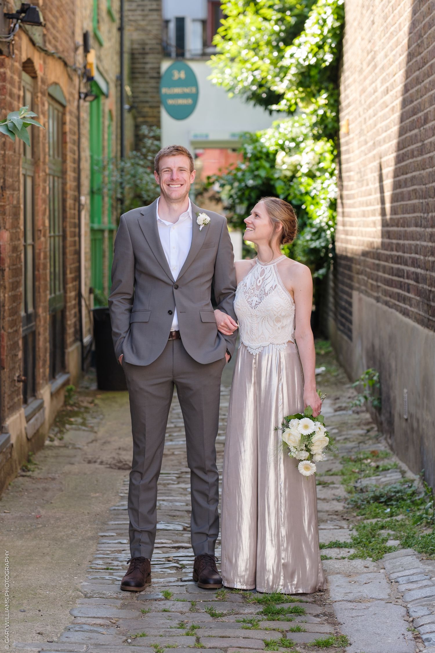 A couple in formal attire poses in a romantic narrow alleyway with brick walls and greenery in the background.