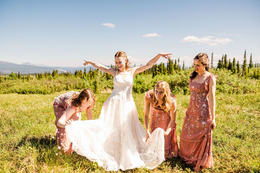 Bridesmaids help bride with dress in mountain meadow during wedding celebration.