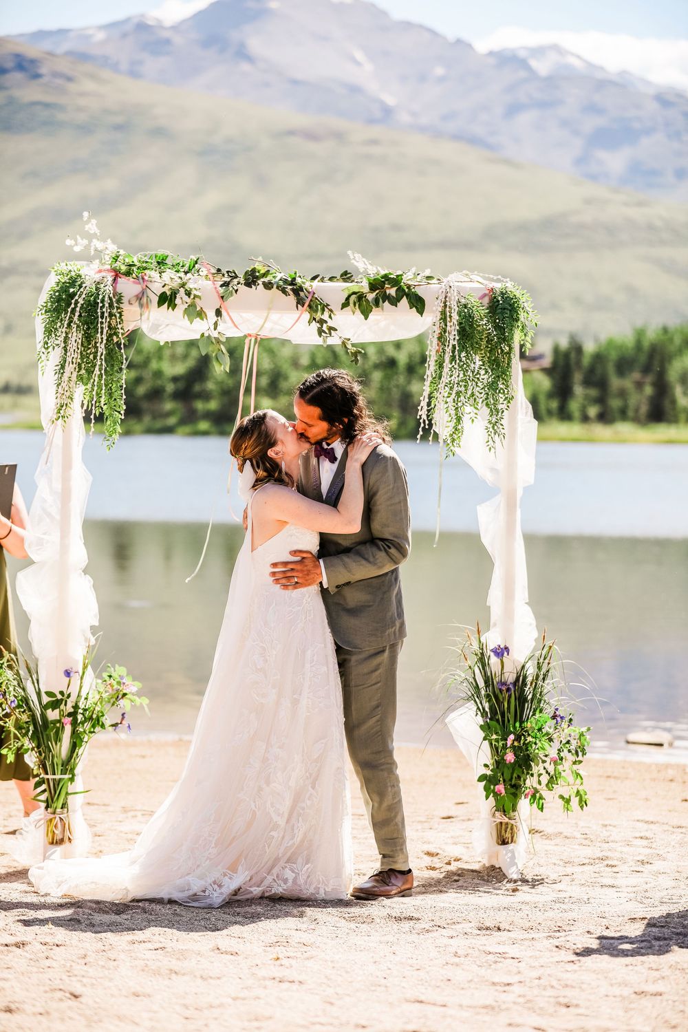 Couple sharing first kiss under white floral arch with mountain and lake backdrop.