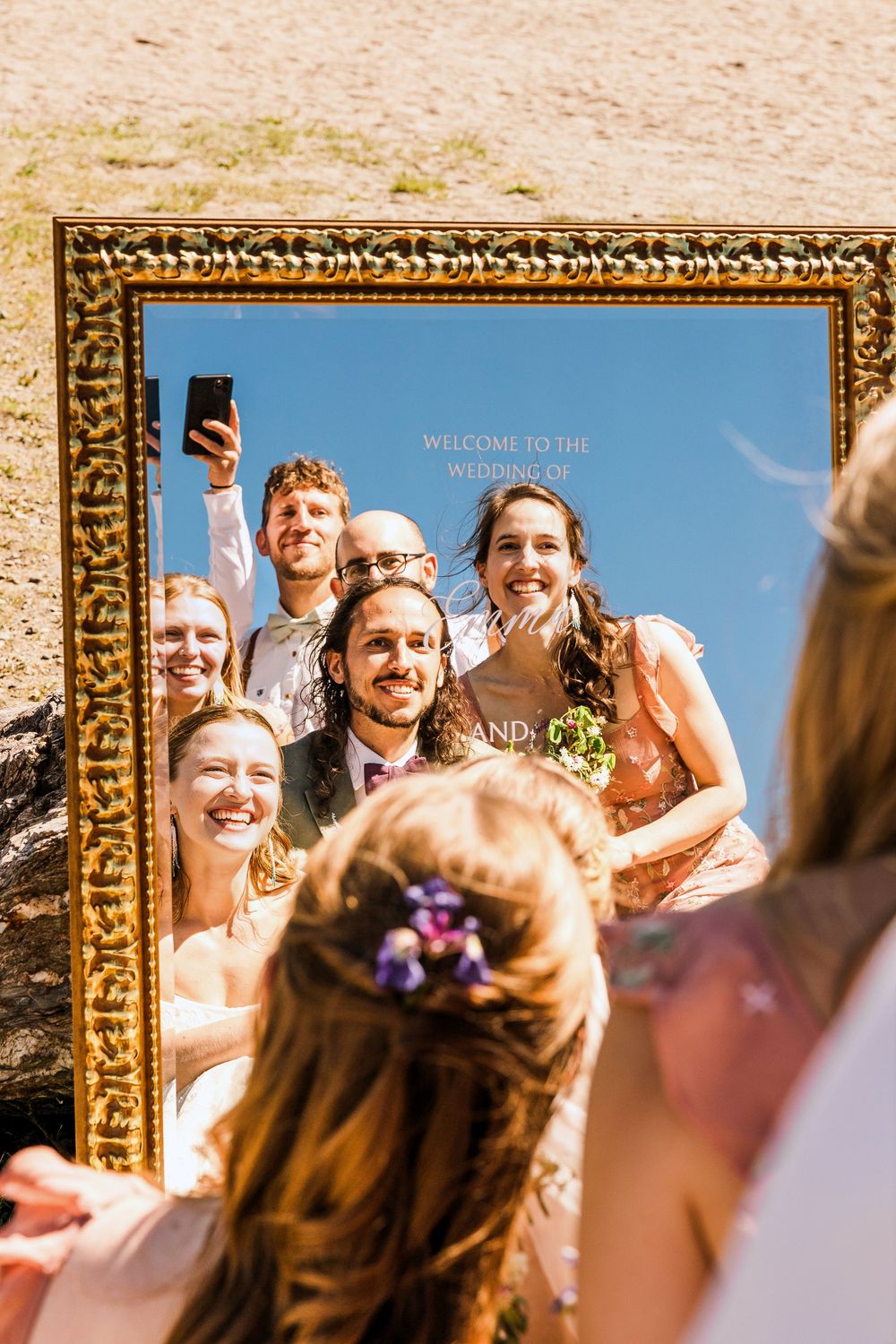 Wedding party taking selfie reflection in ornate gold mirror frame outdoors.