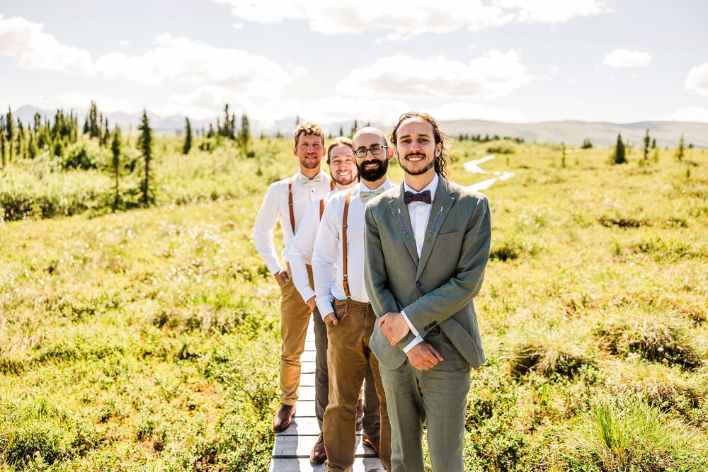 Wedding party lines up on wooden boardwalk against mountainous backdrop with sunny sky.