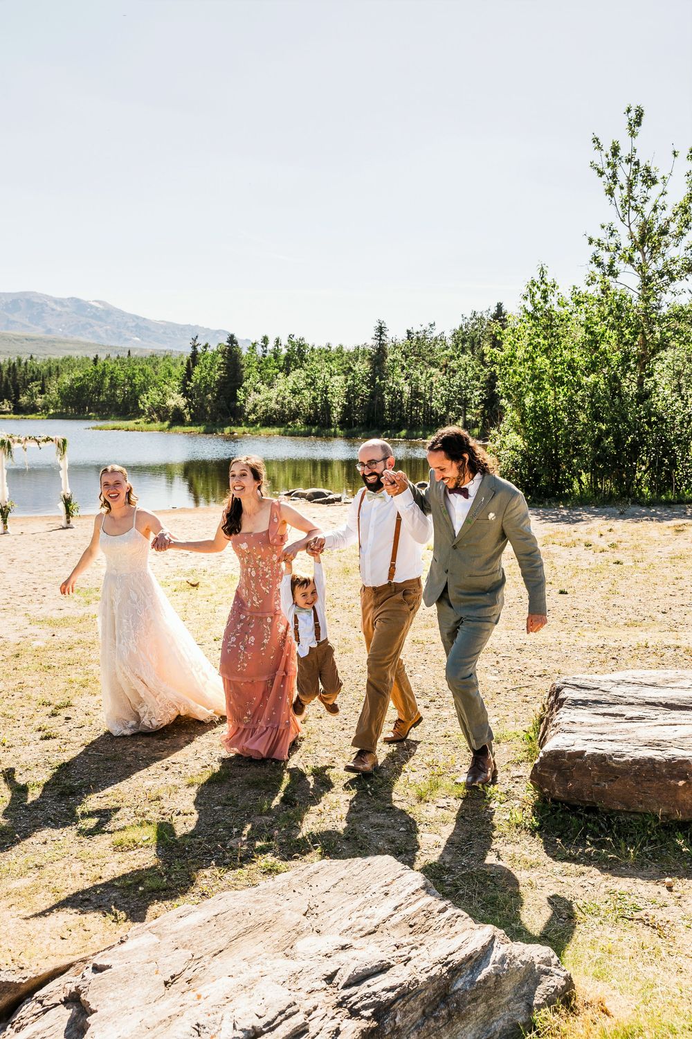Wedding party walks together along a scenic mountain lake on a sunny day.