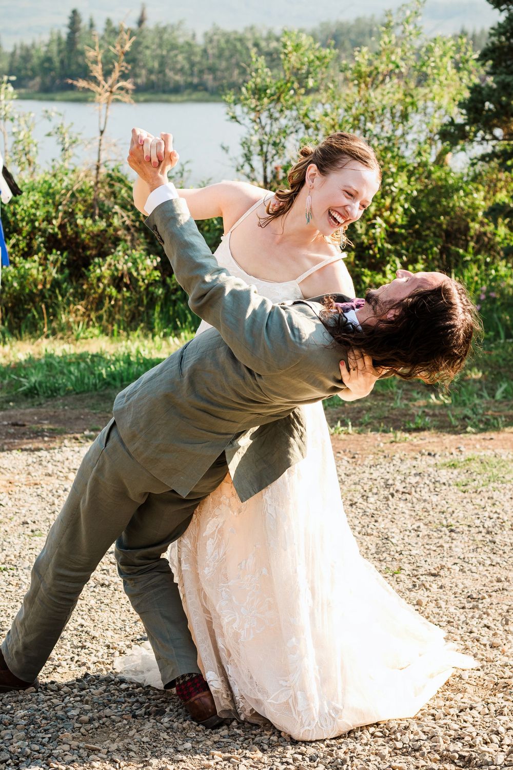 A couple performs a playful dip pose during their wedding photoshoot by a lake.