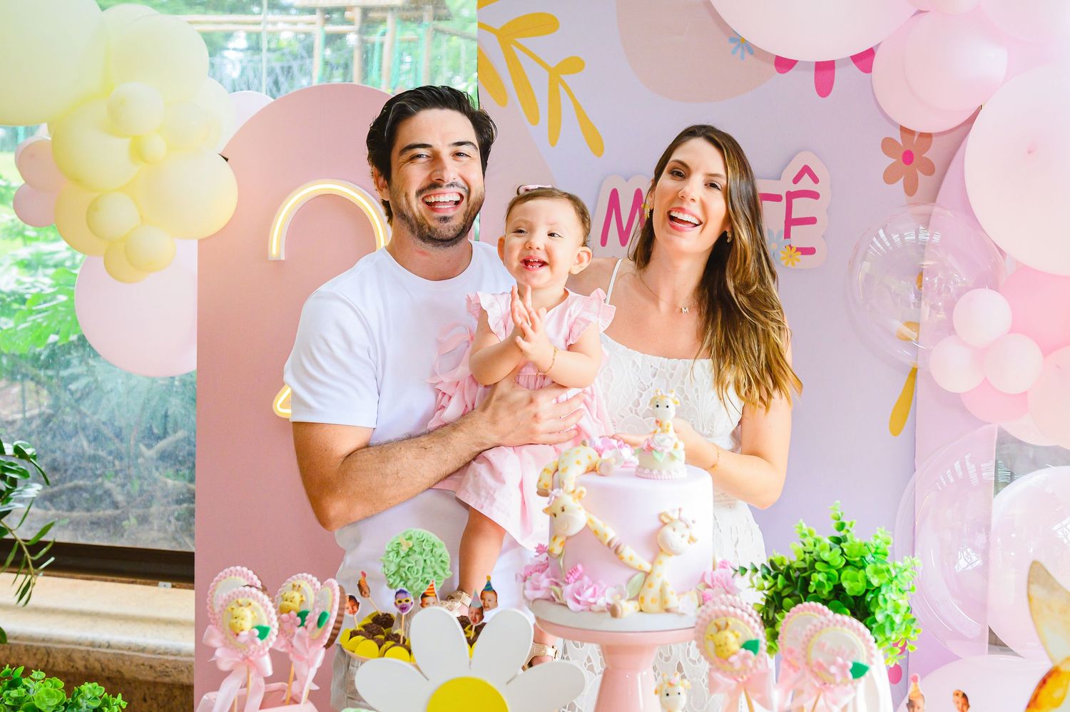 A baby's first birthday celebration with pink and yellow balloon decorations and a decorative pastel cake.