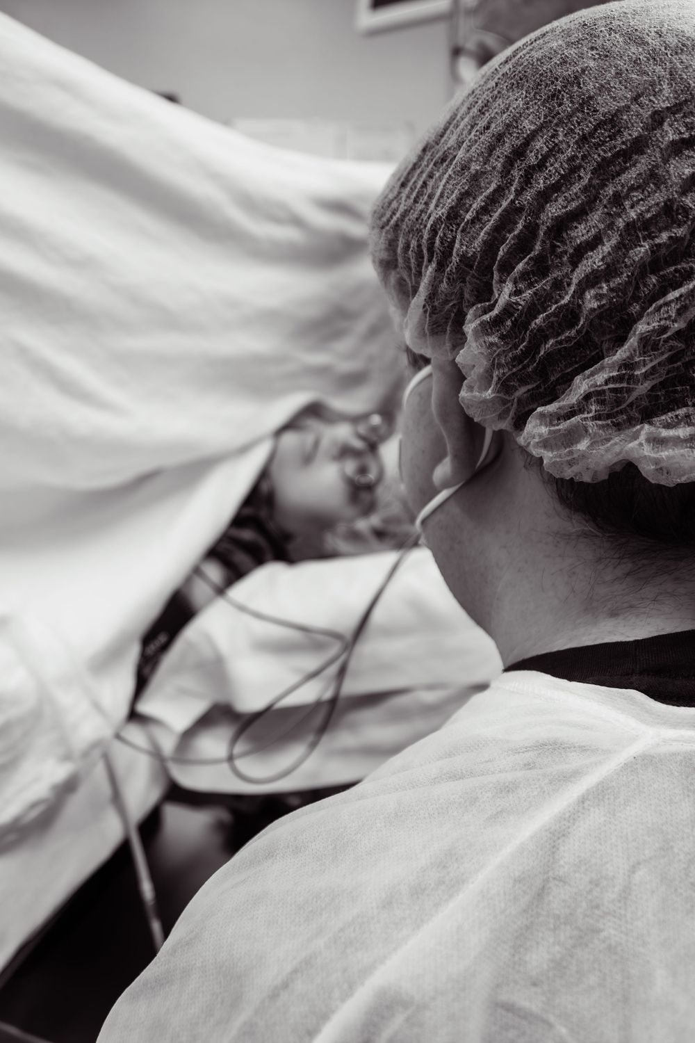 A man wearing a protective hair cap tends to a woman in a medical facility, captured in black and white.