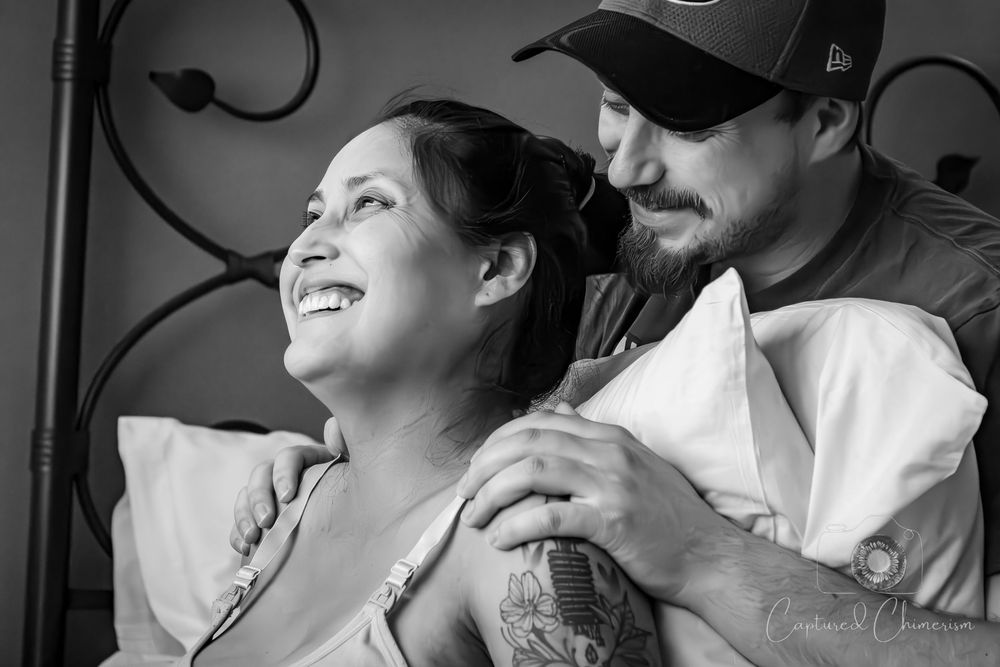 Black and white photo of couple embracing and laughing together on cushions during active labor.