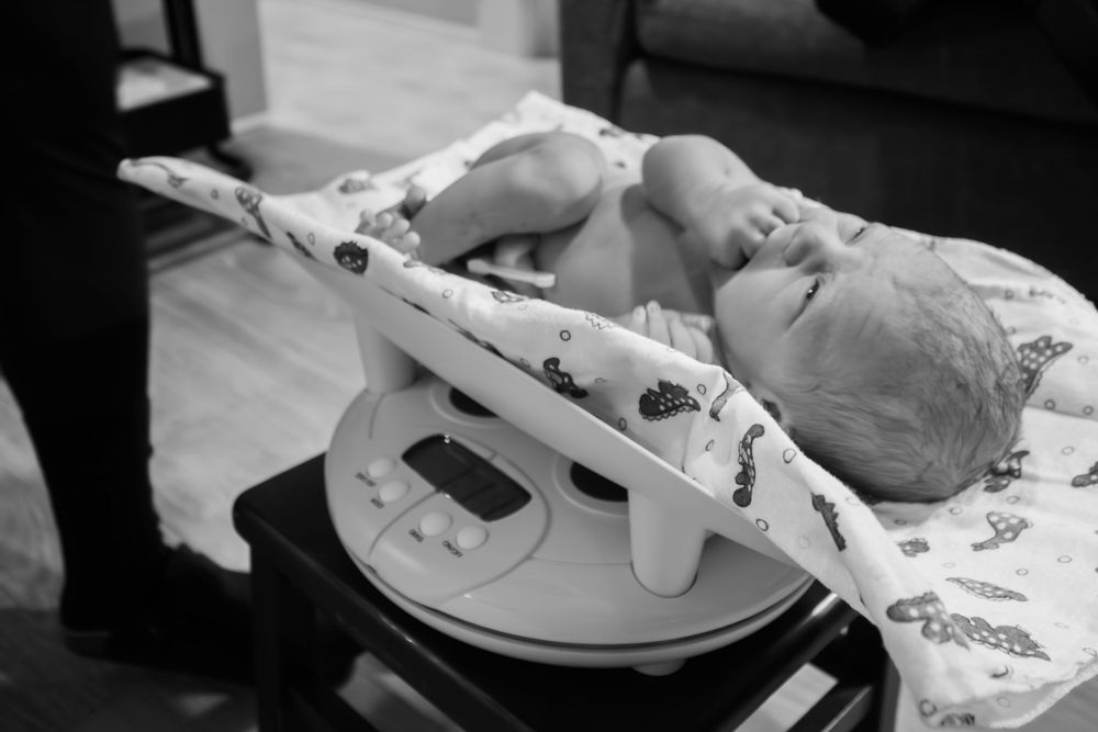 A newborn baby lies on a digital weighing scale during a medical checkup, captured in black and white photography.