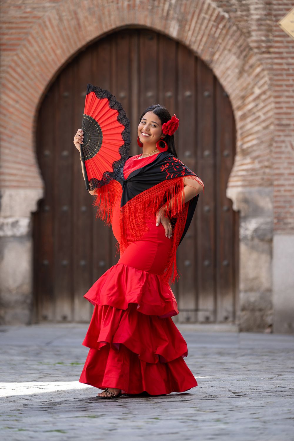 Traditional flamenco dancer in flowing red dress performs with matching fan in front of arched brick doorway.